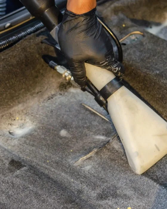 Gloved hand running a hot-water extractor tool over a dirty car carpet